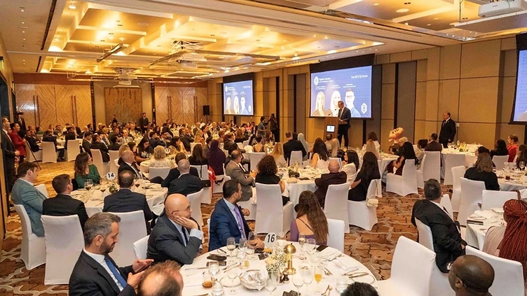 Guests seated at round tables in a chandelier-lit ballroom during the British Chamber of Commerce Qatar Celebration Evening at Park Hyatt Doha, with a speaker at a podium and British Chamber of Commerce Qatar branding on large screens behind the stage.