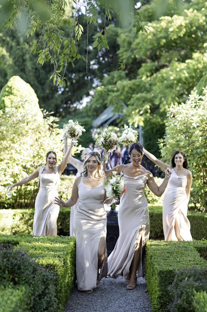 Bridesmaids in champagne satin dresses walking through the gardens at John Joseph Inn in Groton, near Ithaca