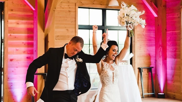 Bride and groom laughing as they walk into their reception with the bride's bouquet raised, surrounded by purple uplighting