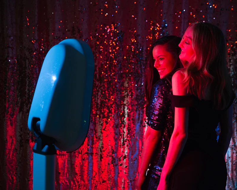 Two guests posing at a modern digital photo booth with a shimmering gold backdrop
