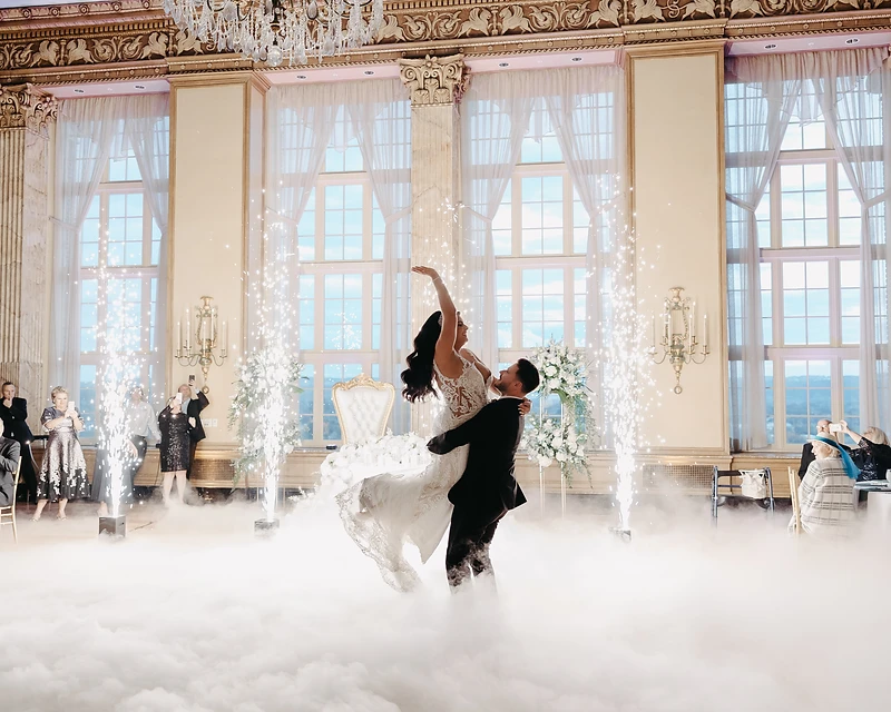 Bride and groom dancing on a cloud of low-lying fog surrounded by four cold spark fountains in a grand ballroom