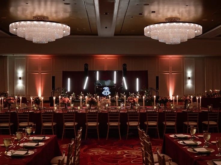 Turning Stone ballroom set for a wedding reception with two tiered crystal chandeliers, vertical LED light tubes behind a DJ booth, red uplighting, and a long head table with taper candles