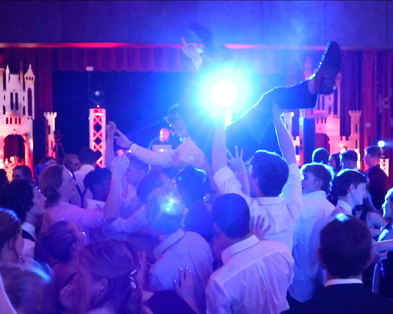 Crowd of students at a high school dance with hands raised under blue and magenta stage lighting