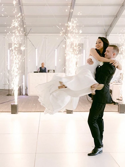 Groom lifting the bride on a white dance floor with four columns of cold sparks inside a wedding tent