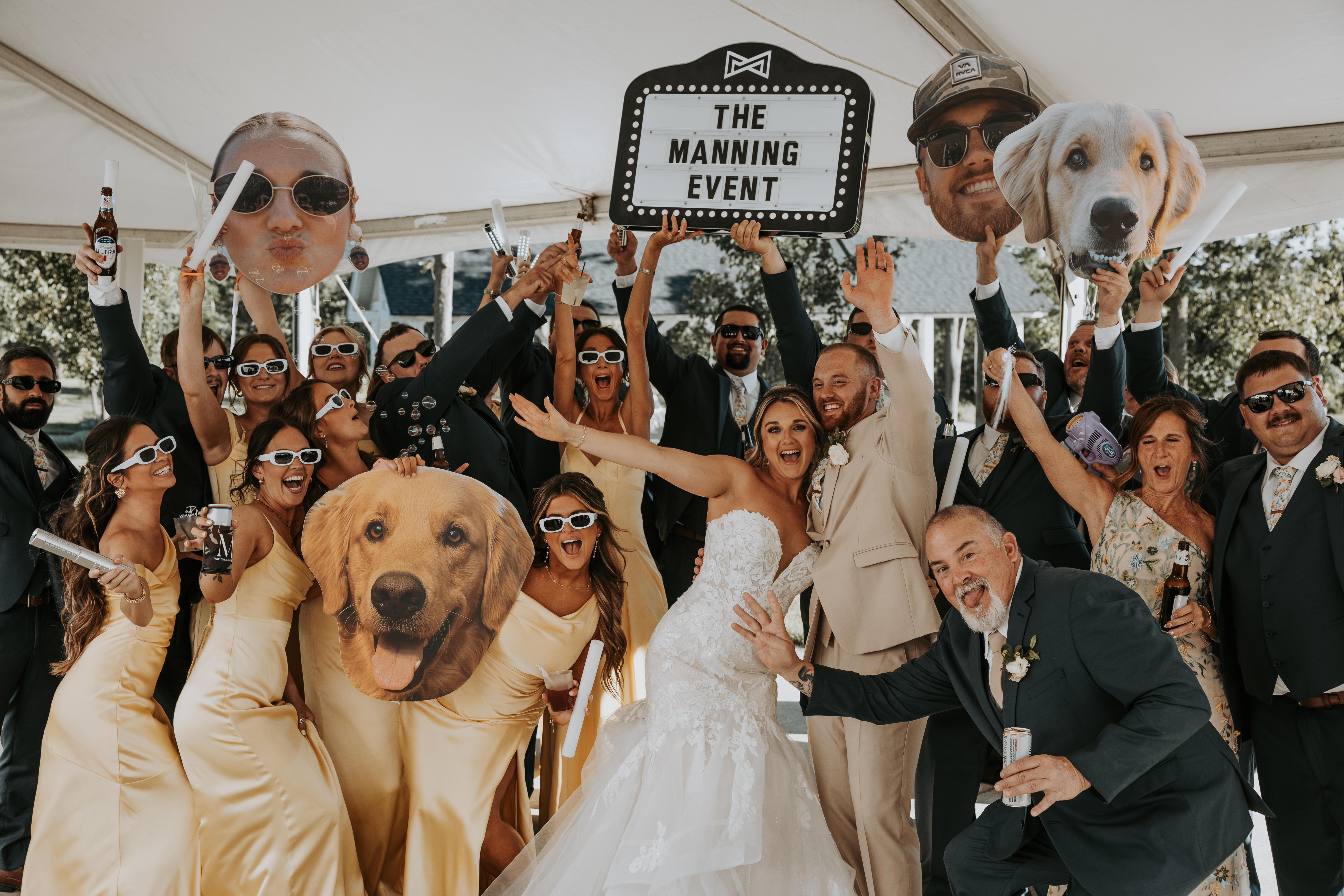 Wedding party holding a custom marquee sign and oversized photo props during a high-energy grand entrance under a tent