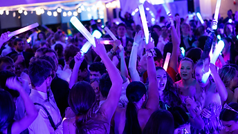 College students with raised glow sticks on a packed formal dance floor under purple and blue lighting