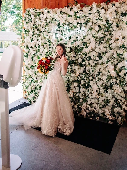 Bride posing with her bouquet in front of a white floral wall with a neon sign at a wedding reception