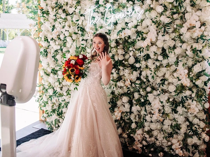 A bride showing her ring in front of an elegant white flower wall with a glowing neon sign