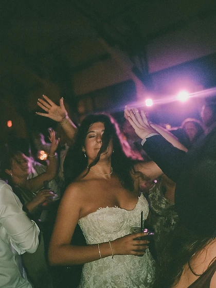 Bride dancing joyfully with arms raised holding a glass on the wedding dance floor