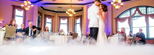 Bride and groom sharing a first dance surrounded by dancing-on-the-clouds fog beneath crystal chandeliers at a Geneva, NY wedding reception