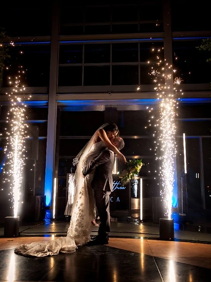 Couple in a dip-kiss framed by two cold spark fountains in a glass atrium with a neon name sign
