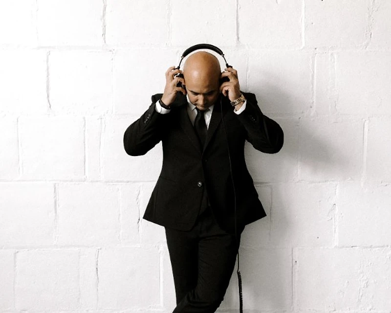 DJ Mike Melice in a black suit and headphones leaning against a white wall