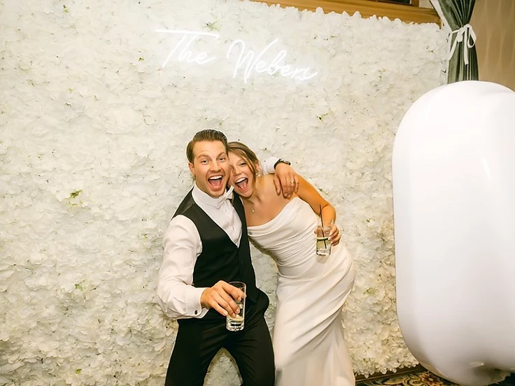 Couple posing at the Social Booth in front of a flower wall with a neon name sign