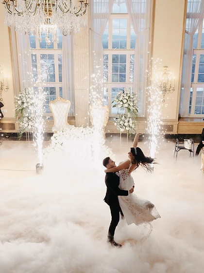 High-angle view of a couple's first dance on low fog framed by four cold spark fountains in a grand ballroom