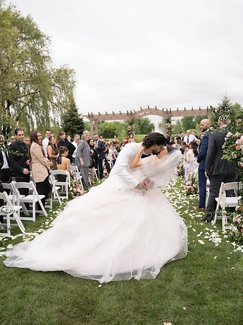 Bride and groom sharing a dip kiss on The Great Lawn at The Lodge, surrounded by white flower petals and guests in white folding chairs at Turning Stone