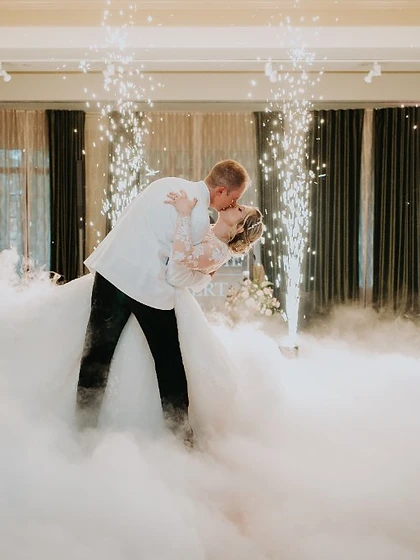 Couple in a dip-kiss on low fog framed by four cold spark fountains at an elegant reception