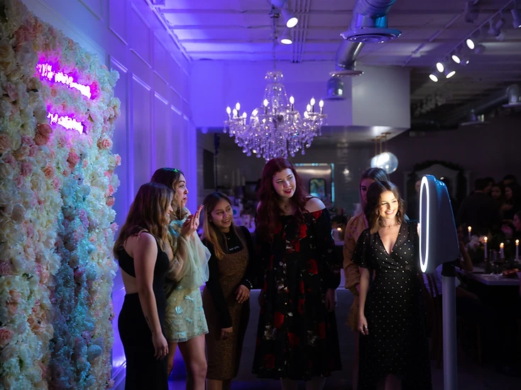 Guests gathered around a modern digital photo booth with a flower wall and neon sign