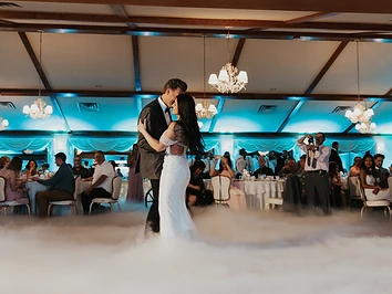 Bride and groom sharing a first dance on a fog-filled dance floor in the Castle Ballroom at Belhurst Castle, with guests watching from candlelit tables and cyan uplighting along the windows