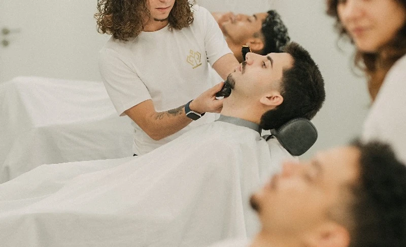 Barbeiro a aparar a barba de um cliente reclinado, ambiente claro e minimalista