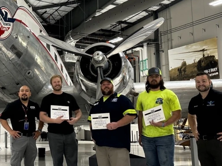 A&P Wash Services aviation crew holding certification certificates inside an aircraft hangar beside a vintage twin-engine plane