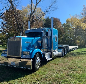 Blue Kenworth semi-truck with flatbed trailer angled on a grassy patch beside a gravel area