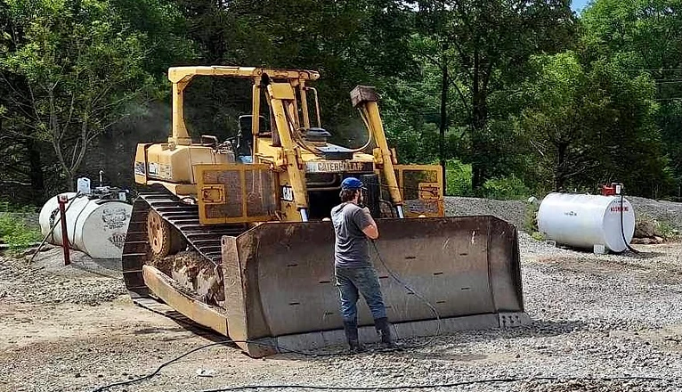 Worker pressure washing a yellow Caterpillar bulldozer in a gravel lot