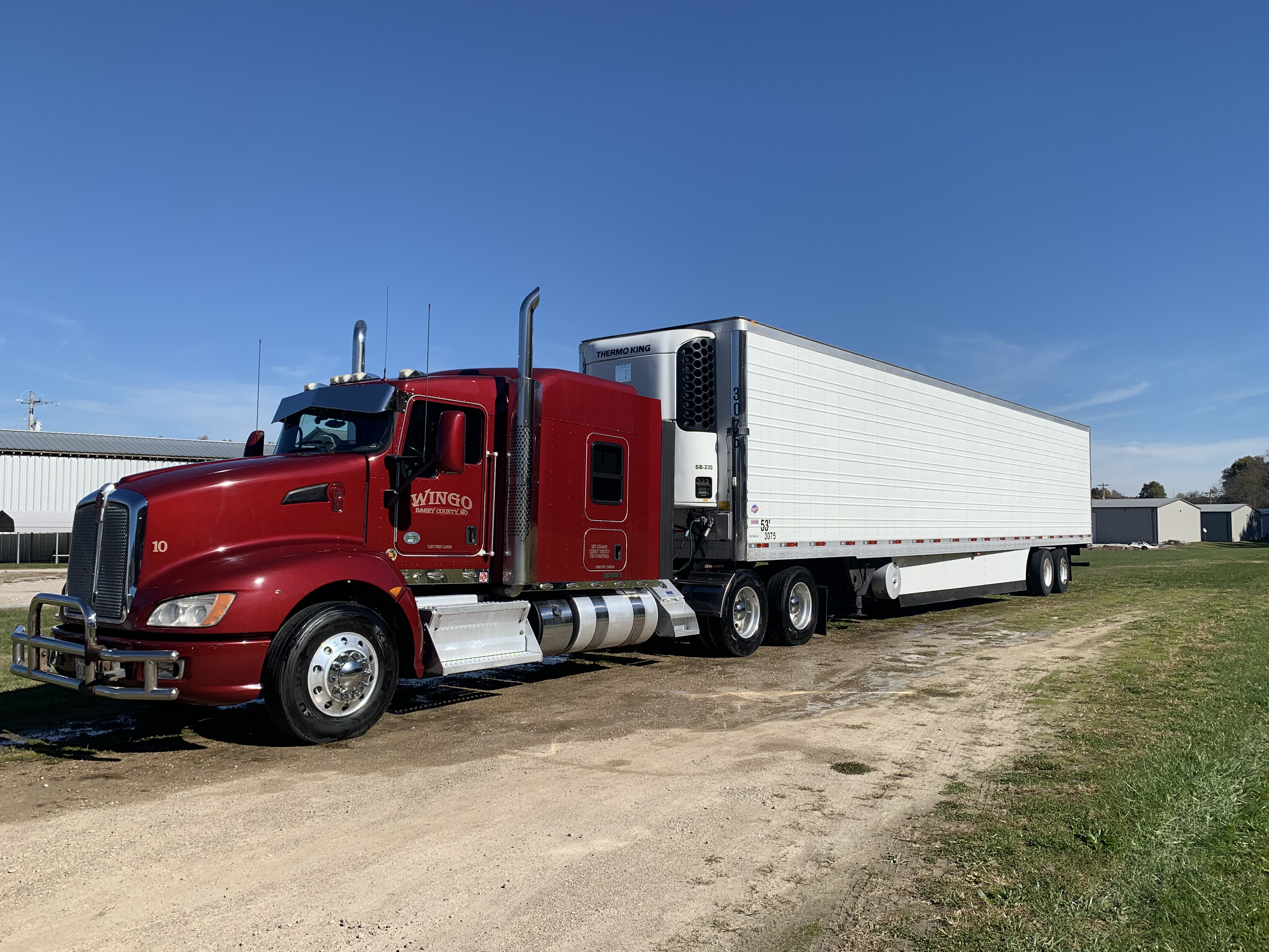 Red Kenworth semi-truck with a white Thermo King refrigerated trailer parked on a gravel lot