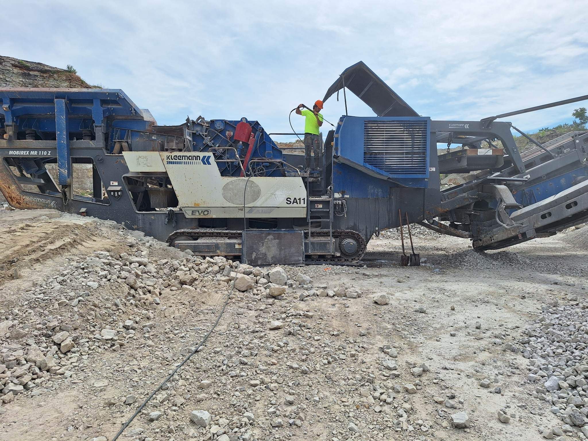 Worker in a high-visibility shirt and hard hat pressure washing a large blue Kleemann rock-crushing machine