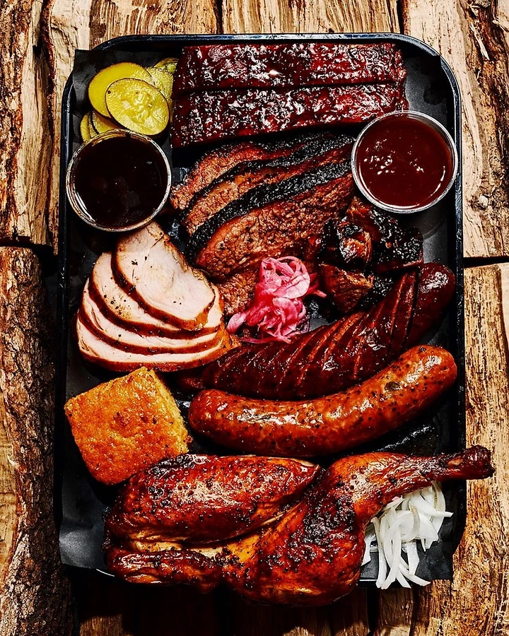 Smoked barbecue platter of ribs, brisket, turkey, chicken, sausage, cornbread and pickles on a black tray over split firewood