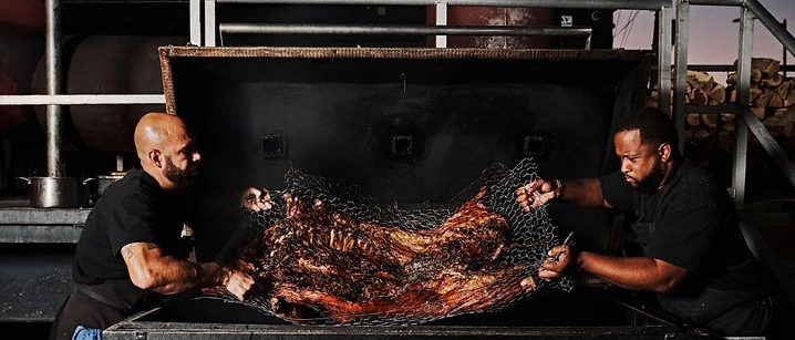 Two chefs lifting a whole smoked hog out of the smoker at dusk