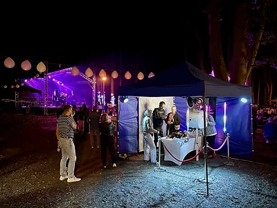 Wieczorne stanowisko fotobudki klasycznej MegaFota pod niebieskim namiotem — podświetlane tuby LED, baner i scena w tle