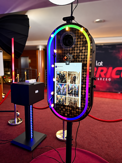 Fotobudka AI MegaFota z galerią portretów AI na ekranie, obok czarna konsola i lampa błyskowa, na czerwonym dywanie przed tłem z logo BRICO i MegaFota