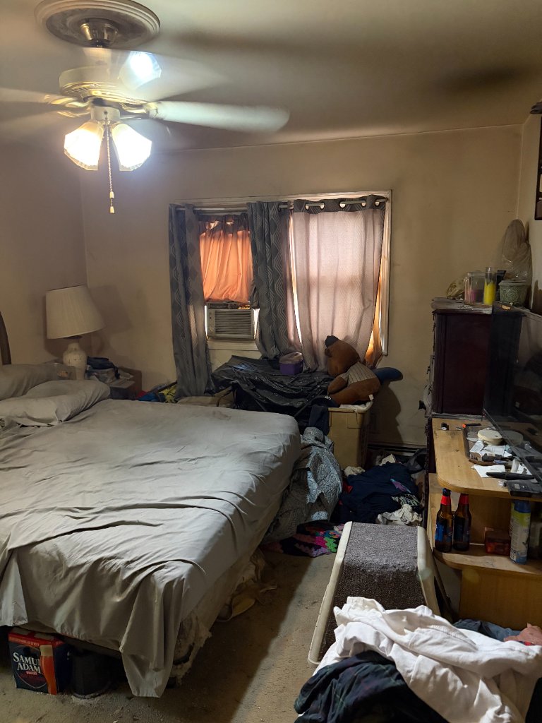 Cluttered bedroom packed with a bed, clothing piles, furniture, and household items before a full clean out