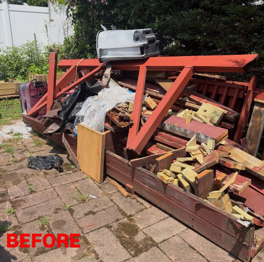 Collapsed red wooden deck broken into framing, boards, and debris piled on a brick paver patio before removal