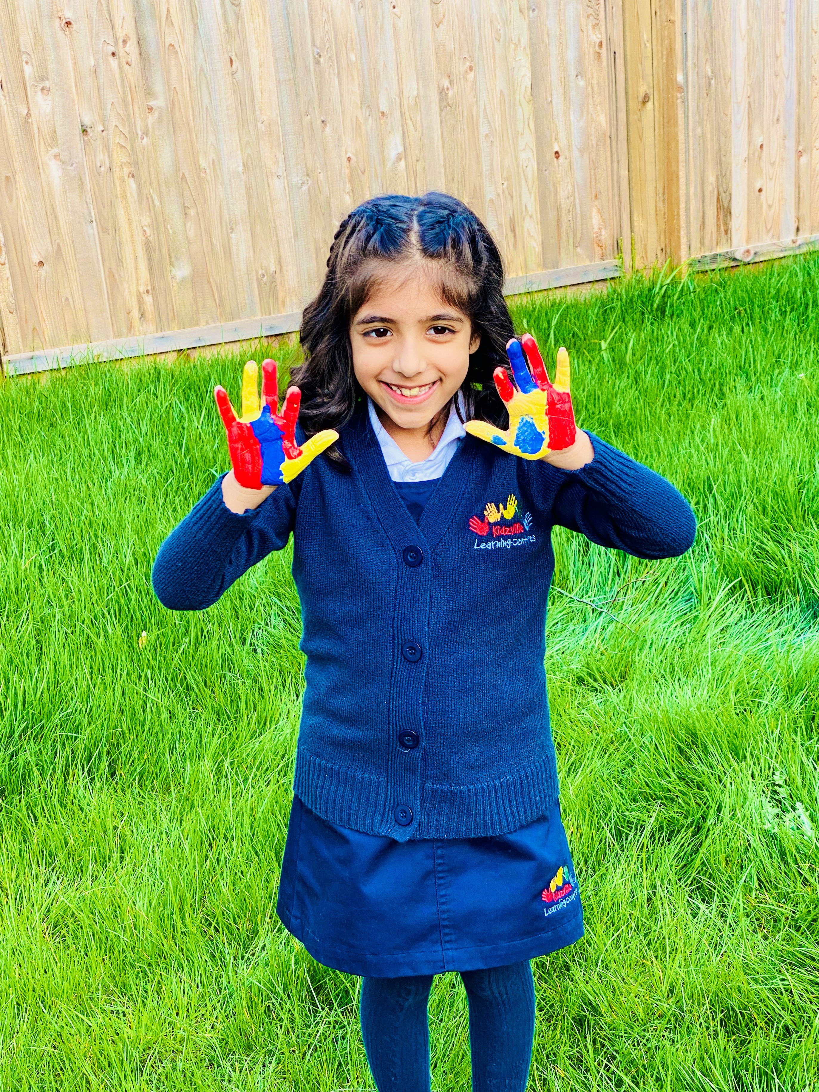 A smiling Kidzville student in uniform holding up paint-covered hands outdoors