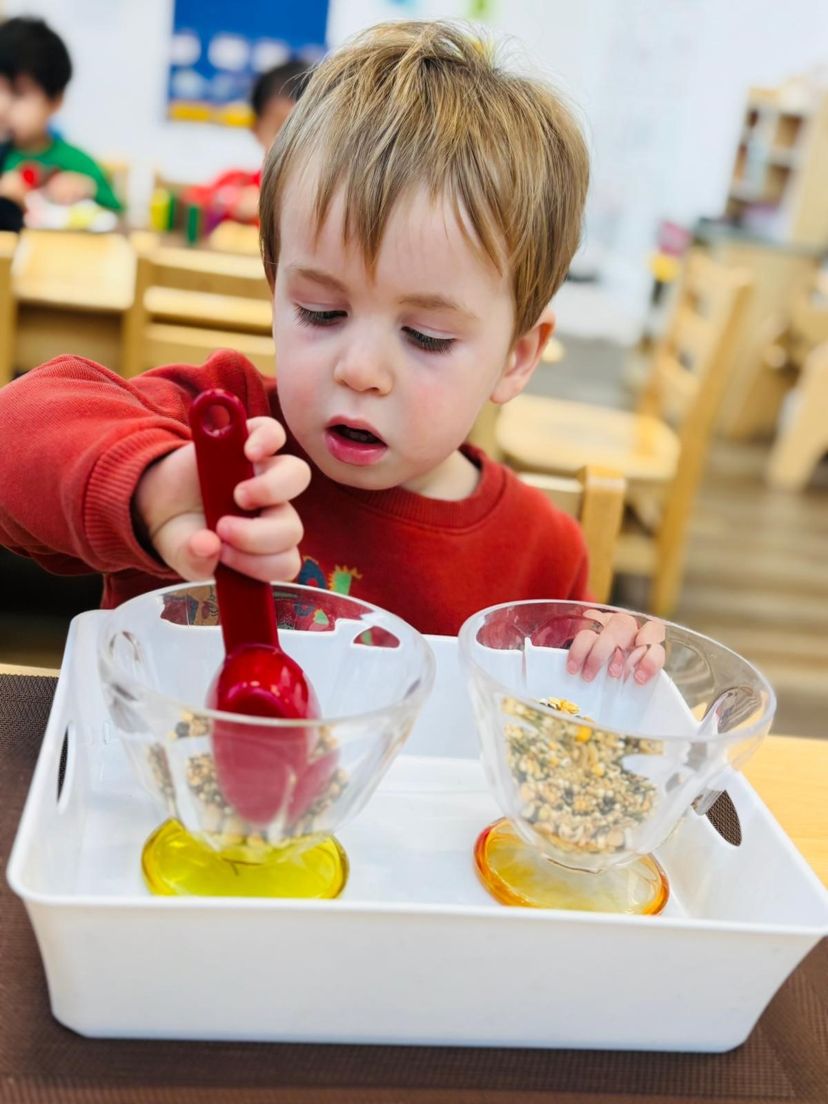 Toddler using tongs to transfer seeds between glasses at Kidzville