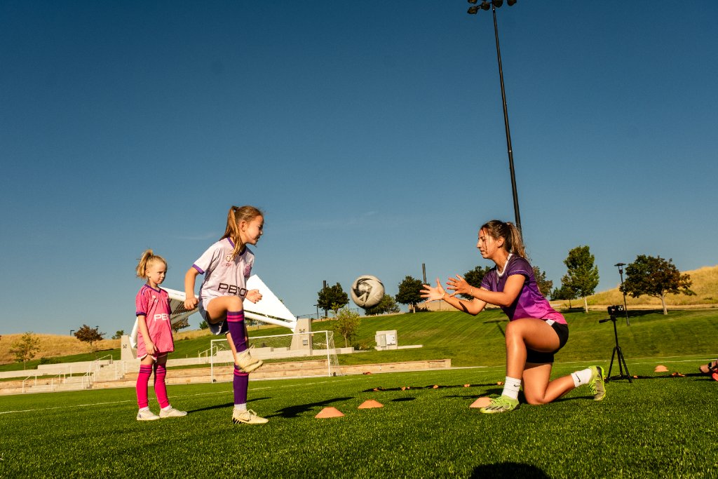 Coach Ana training a small group of young female soccer athletes together on a sunny field