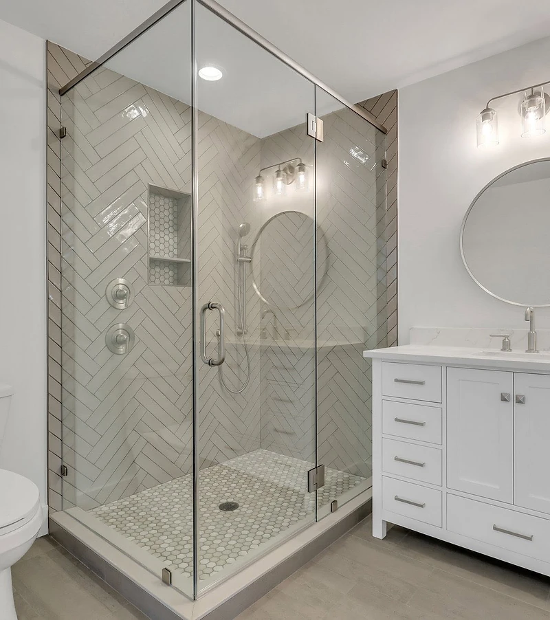 Herringbone tile corner shower with glass enclosure, hexagon mosaic floor, and white vanity