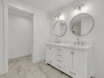 Double vanity with two round mirrors, marble-look floor tile, and pocket door to water closet