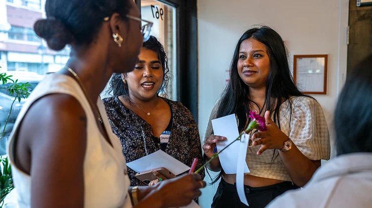 Three women in conversation at an Empower'em gathering, one holding a flower with a quote tag