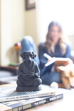 A dog Buddha figurine in sharp focus atop copies of My Dog, My Buddha, with the author writing and a dog softly out of focus in the background.