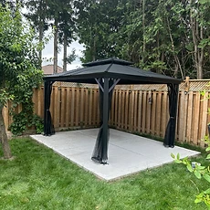 A completed black metal gazebo stands on a concrete patio next to a wooden fence.