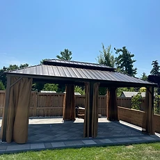 A metal-roofed gazebo stands on a stone patio with curtains closed.