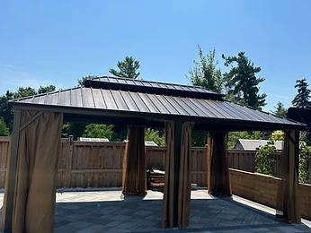 A metal-roofed gazebo stands on a stone patio with curtains closed.