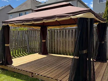 A completed gazebo with brown fabric canopy and curtains on a wooden deck in a backyard.