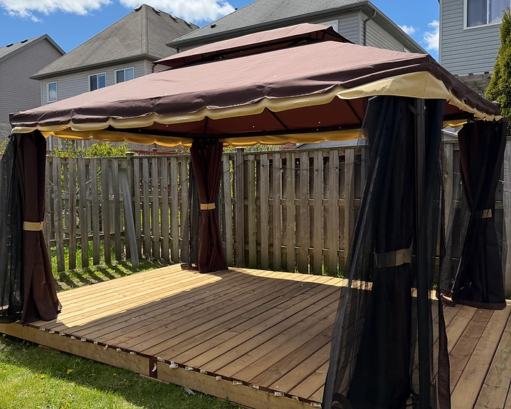 A completed gazebo with brown fabric canopy and curtains on a wooden deck in a backyard.