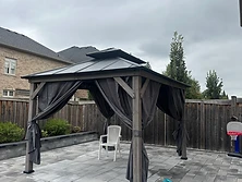 A gray gazebo with curtains stands on a stone patio in a backyard.