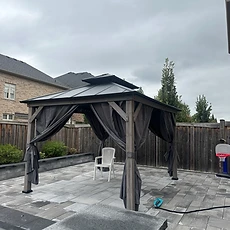 A gray gazebo with curtains stands on a stone patio in a backyard.