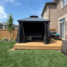 A fully assembled metal gazebo stands on a wooden deck next to a residential house.