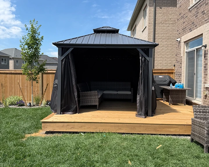 A fully assembled metal gazebo stands on a wooden deck next to a residential house.